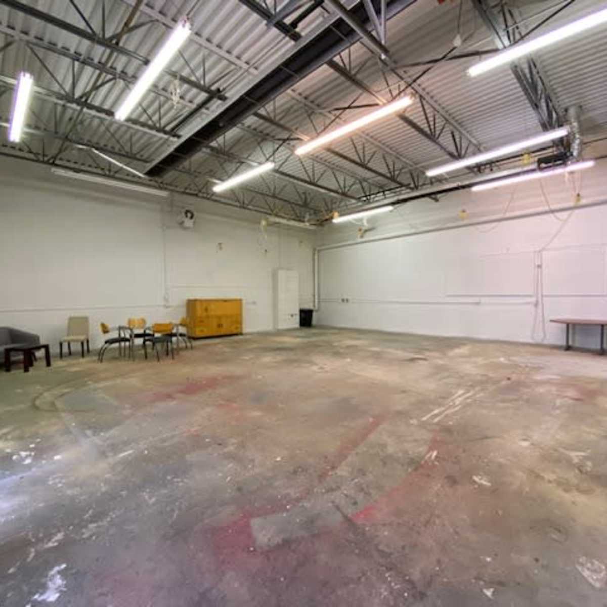 An empty industrial space features a concrete floor, scattered furniture, and overhead lighting.