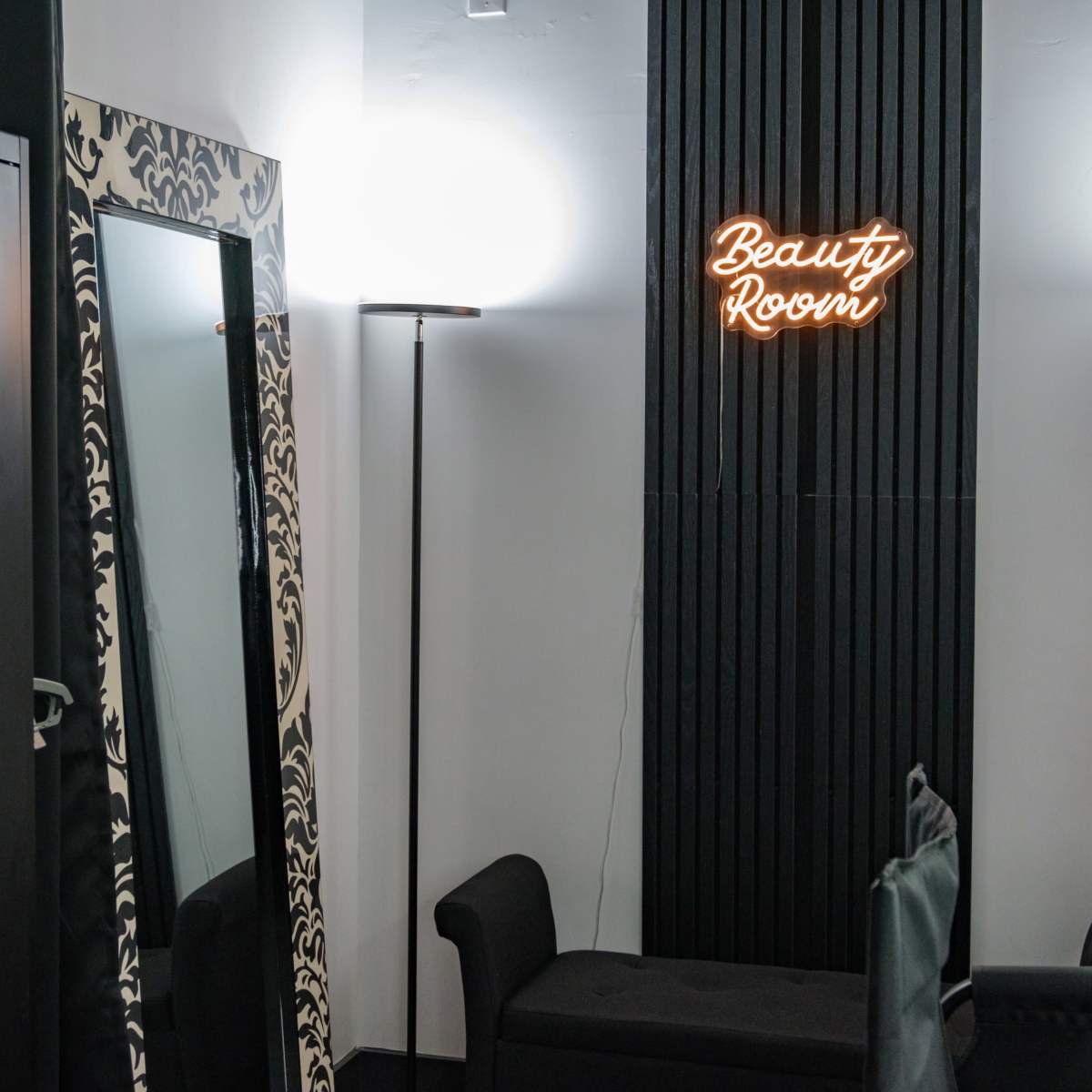 A beauty room with a large mirror, a floor lamp, and a neon sign reading "Beauty Room" on the wall.