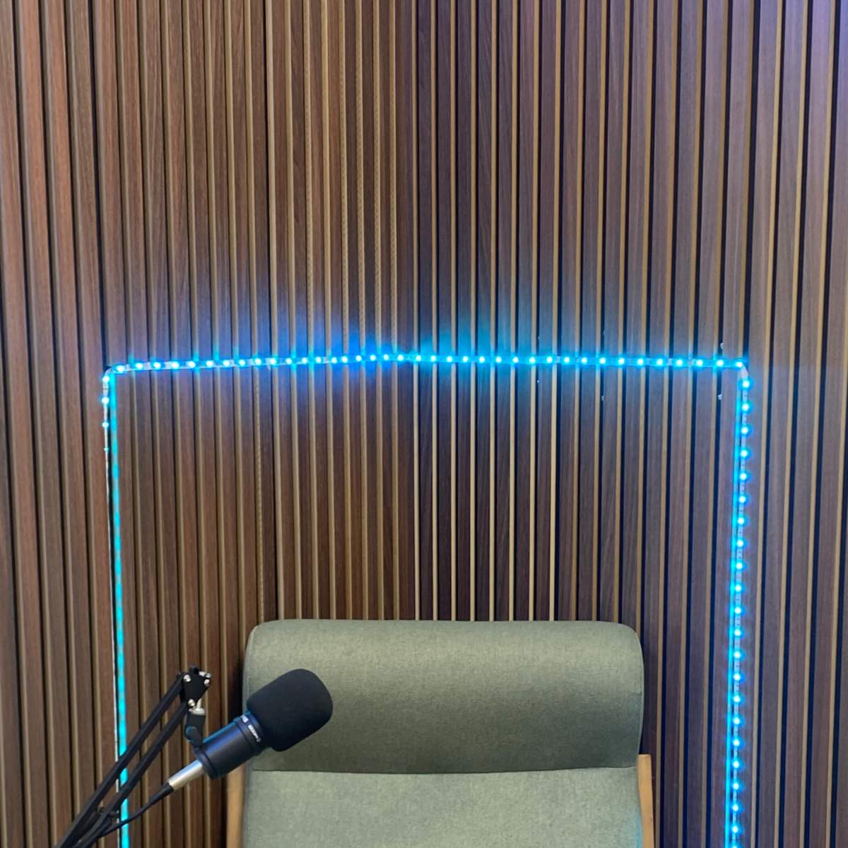 A lighted rectangular frame outlines a green chair beside a microphone and wooden table against a wooden slat wall.