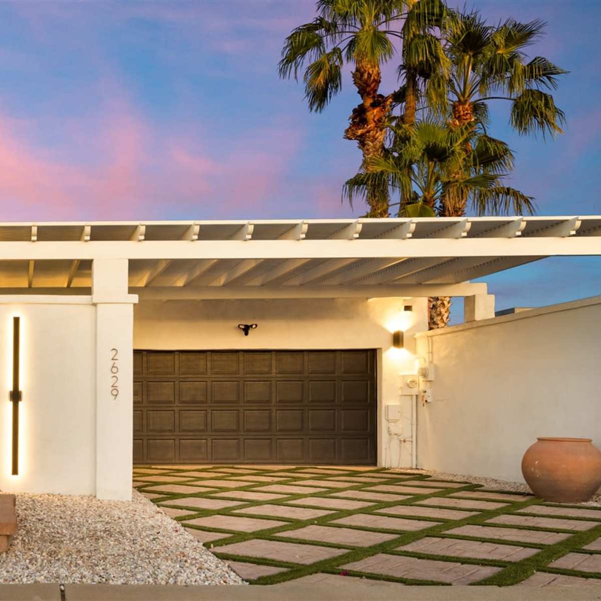 A modern home features a minimalist design with a gray garage door, a stone pathway, and a palm tree against a colorful sunset sky.