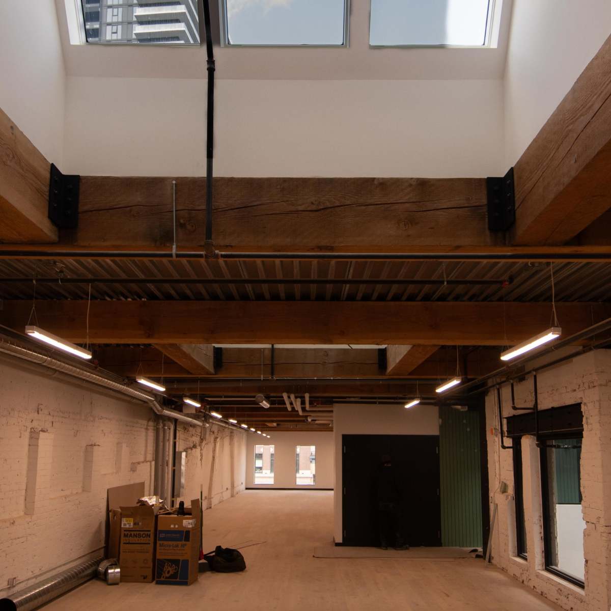 An empty industrial-style room with high ceilings, wooden beams, and skylights, featuring cardboard boxes and a dark doorway at one end.
