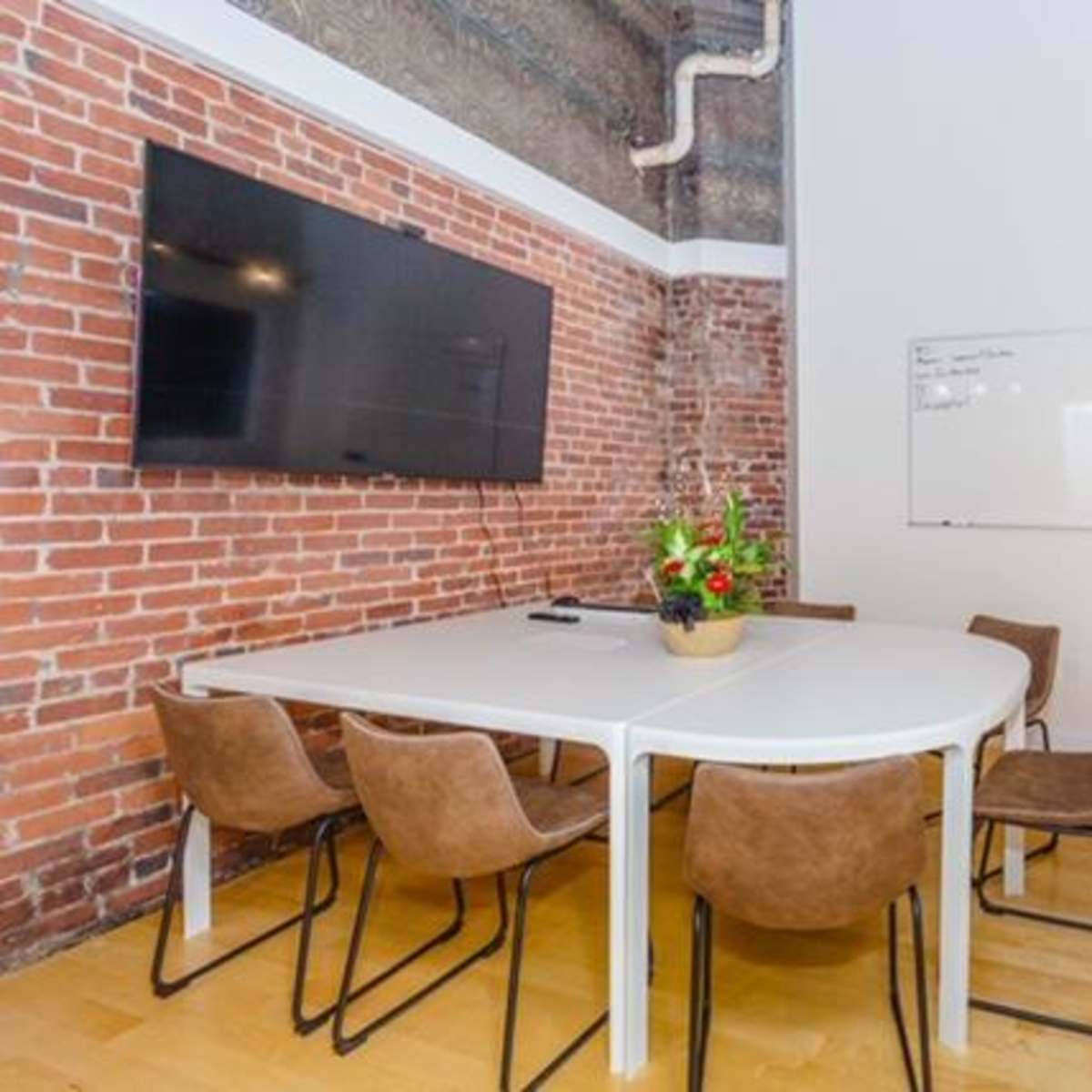 A conference room features a round table surrounded by brown chairs, a large wall-mounted TV, and a whiteboard on the adjacent wall, all set against a backdrop of exposed brick.