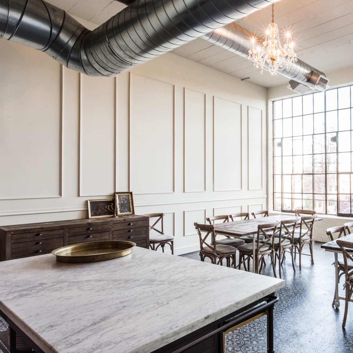 A spacious room with large windows, featuring a marble table at the center, multiple wooden dining tables arranged throughout, and a decorative chandelier hanging from the ceiling.