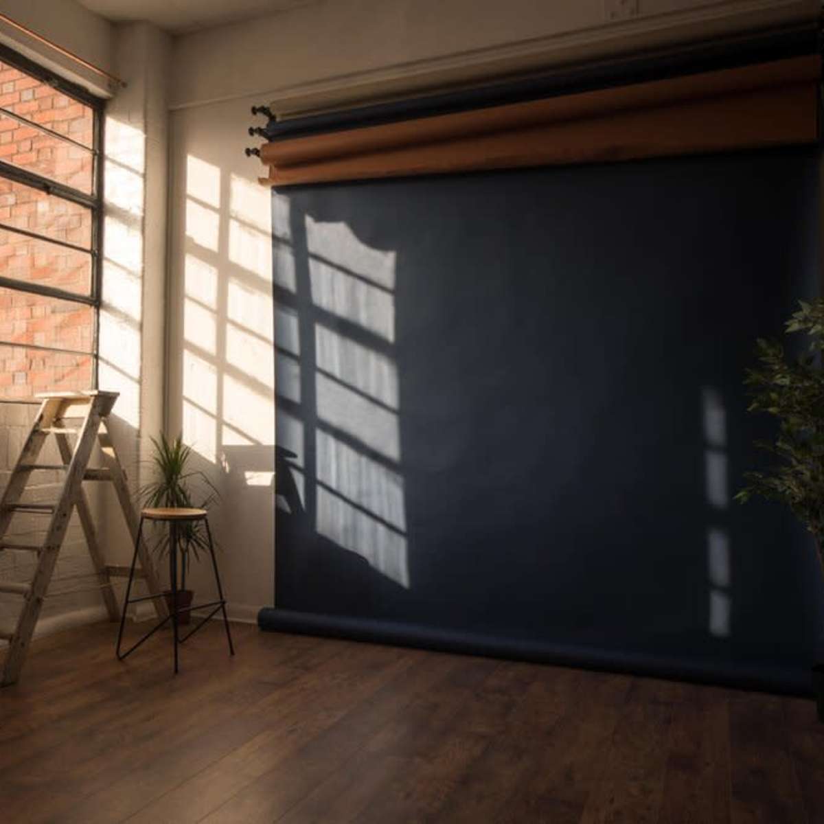 A spacious interior with a large window, a wooden ladder, a stool, a potted plant, and a dark backdrop rolled down against a wall.