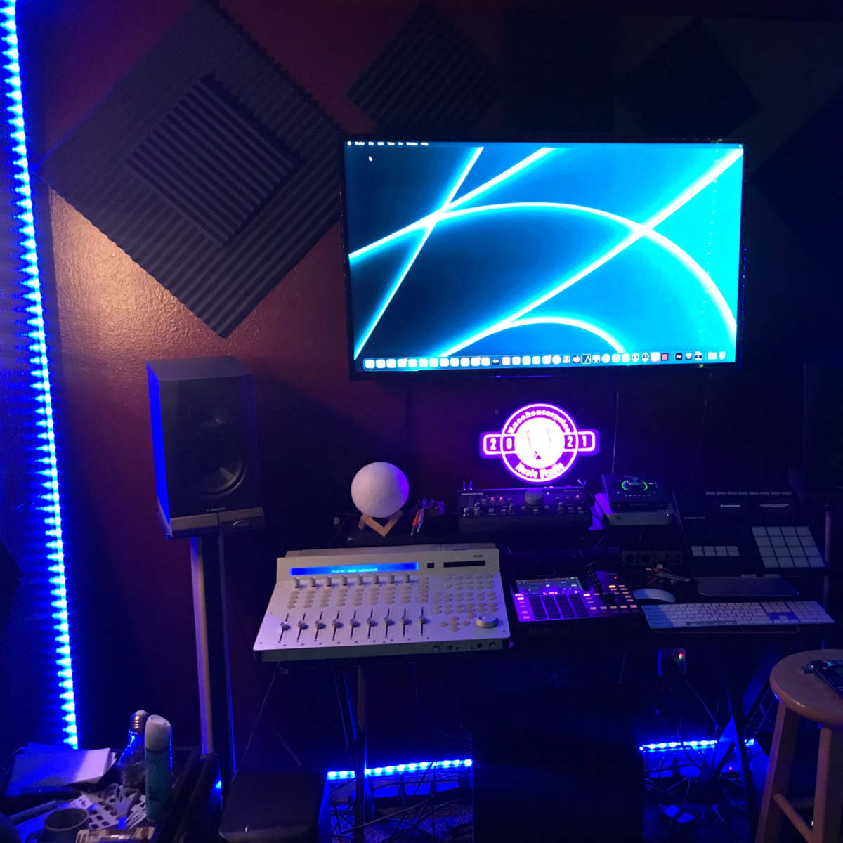 A music production studio features a computer screen displaying blue graphics, various electronic instruments, and ambient lighting.