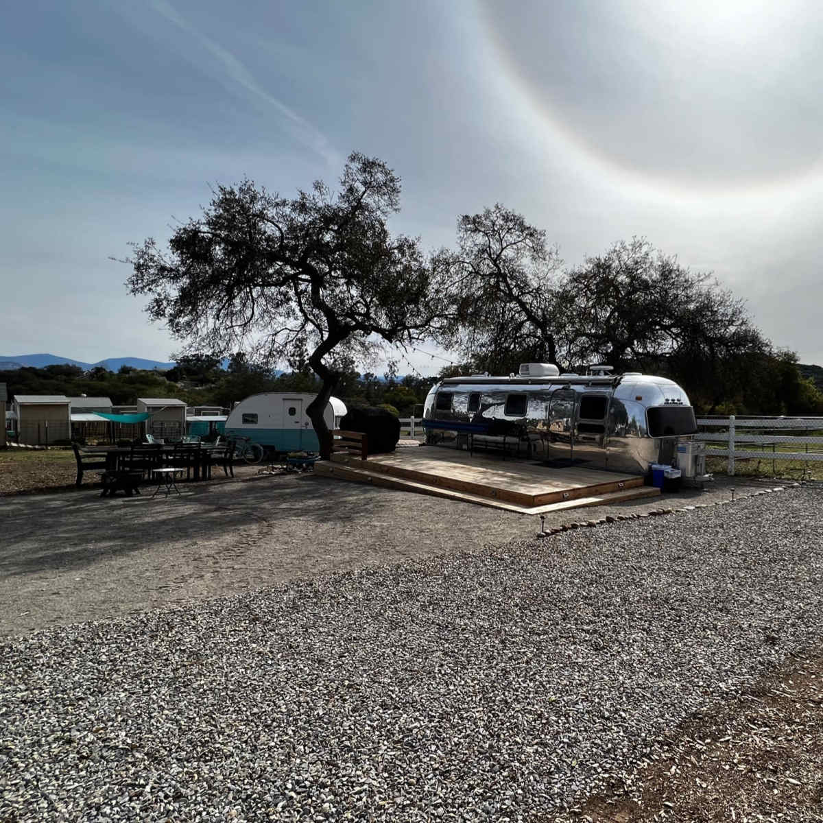 An Airstream trailer is parked on a gravel area under a large tree, with a wooden deck and a fenced field in the background.
