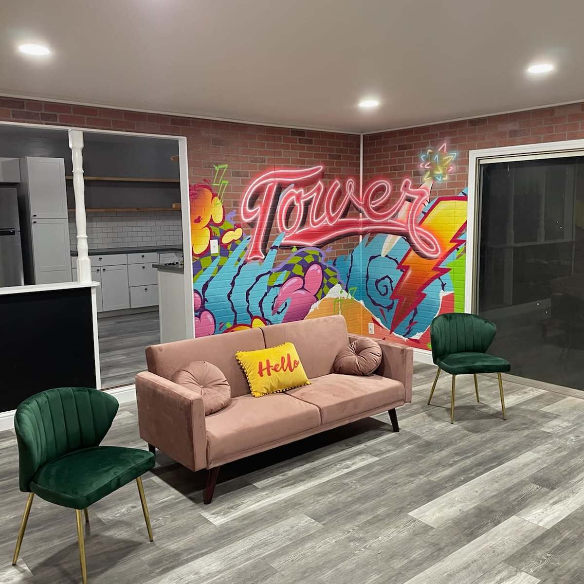 A modern living space features a pink sofa with a "Hello" pillow, two green chairs, and a colorful mural reading "Tower" on the wall.