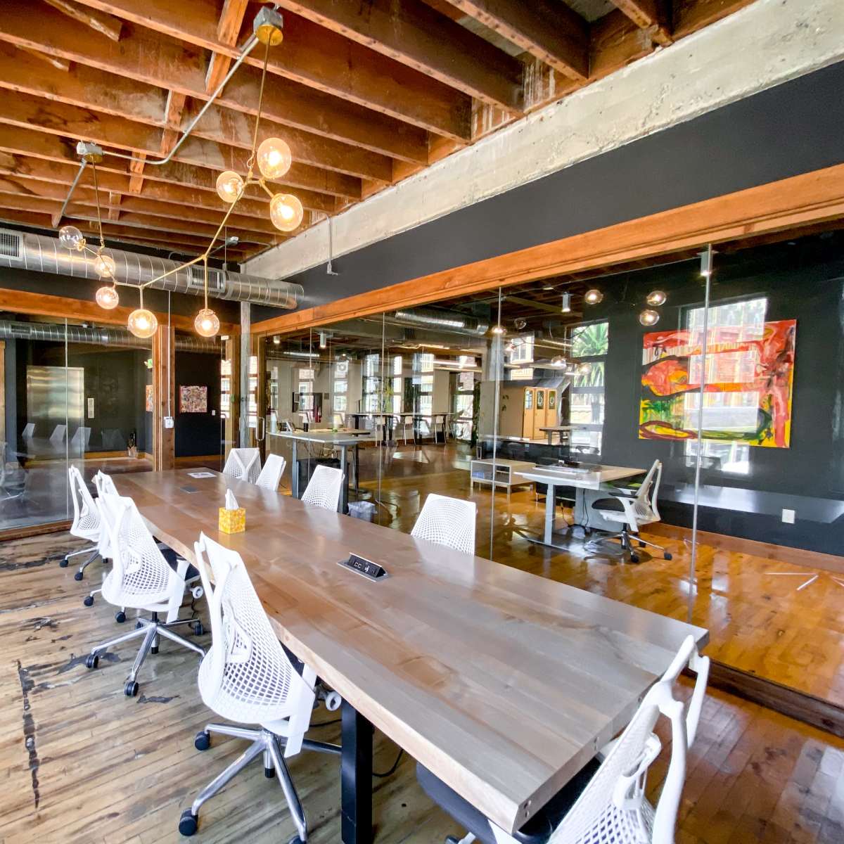 A large, modern conference room features a long wooden table surrounded by white ergonomic chairs, with exposed wooden beams and glass walls.
