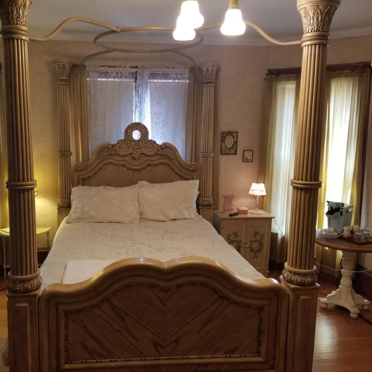 A vintage-style bedroom featuring a large, ornate bed with decorative pillars, soft lighting, and large windows covered with sheer curtains.