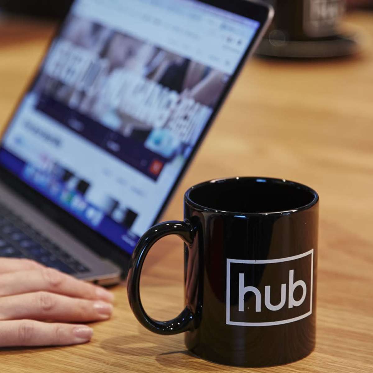 A person uses a laptop on a wooden table with a black coffee mug featuring the logo "hub" nearby.