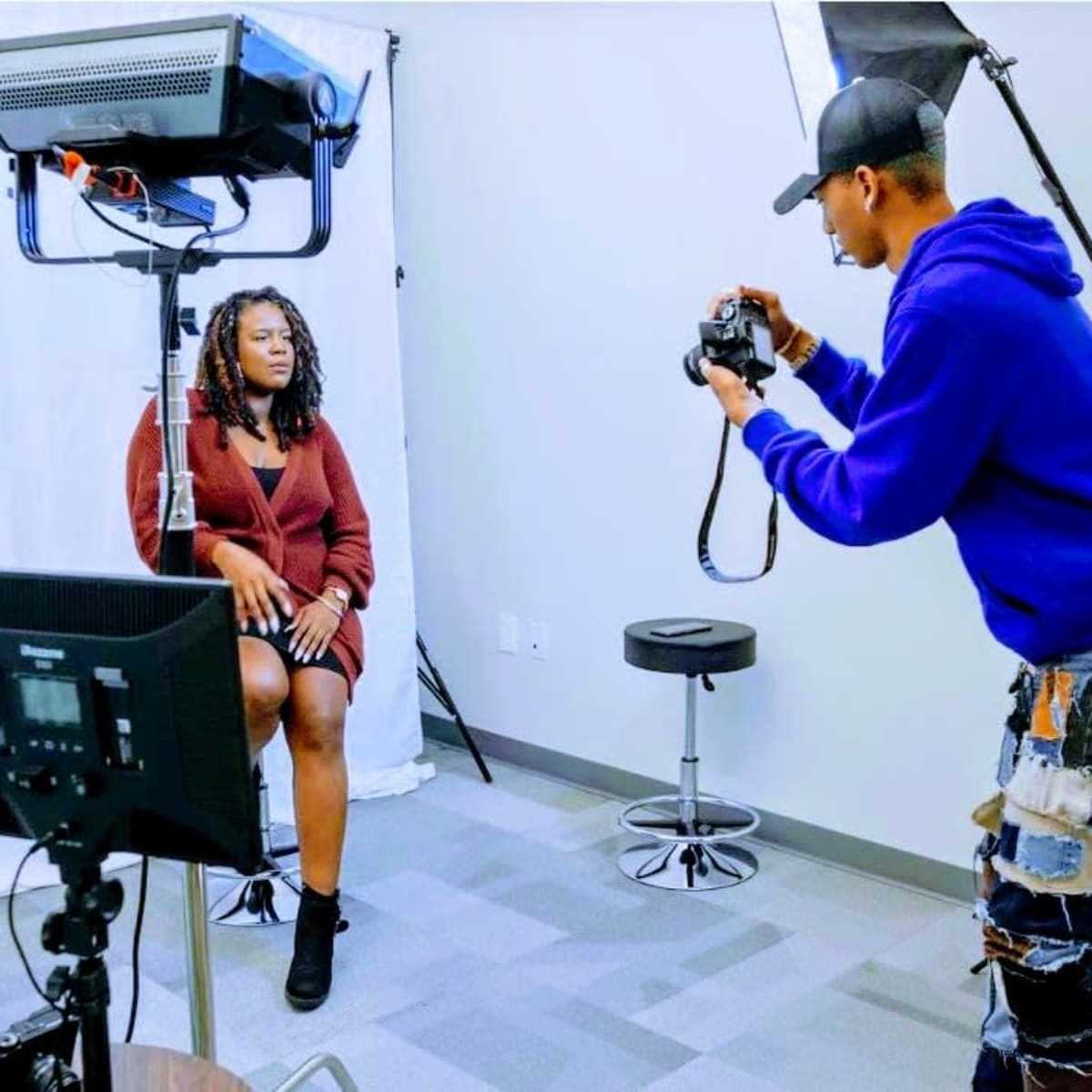 A photographer captures images of a woman seated in a studio setting with lighting equipment and a backdrop.