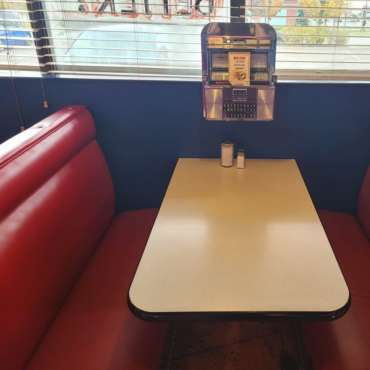 A red vinyl booth with a white tabletop is set up in a diner, featuring a vintage jukebox and condiments on the table.