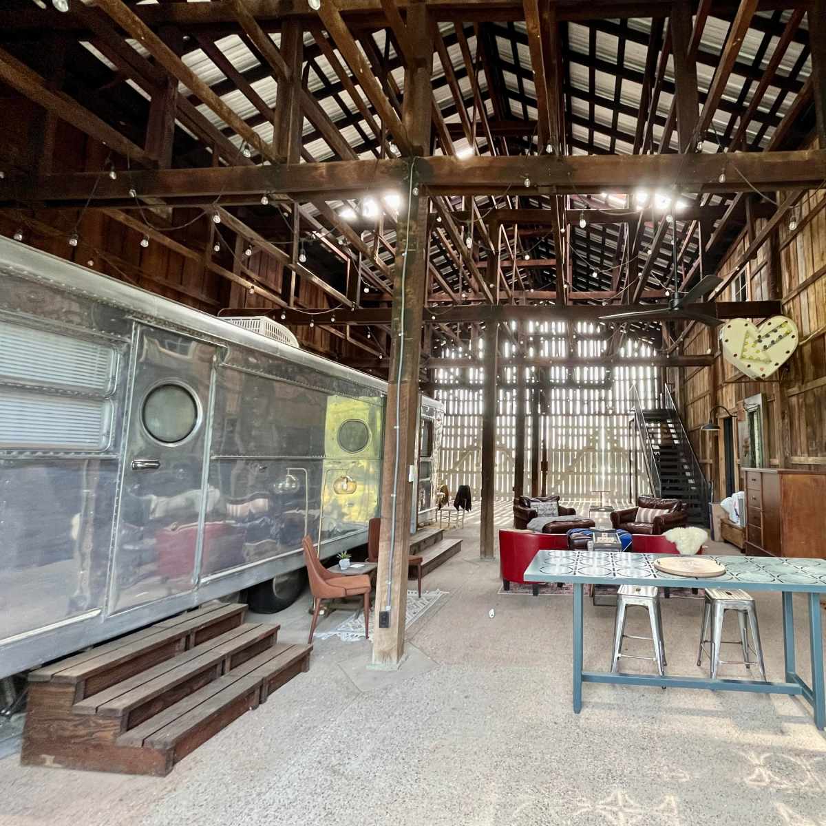 A spacious barn interior with a vintage trailer on one side, a dining table, and lounge seating arranged in an open layout.
