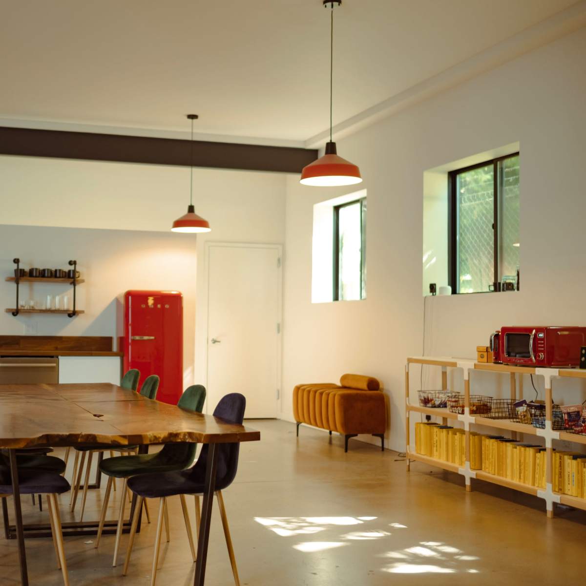 A modern kitchen and dining area features a large wooden table surrounded by colorful chairs, a red refrigerator, and various appliances on a countertop.