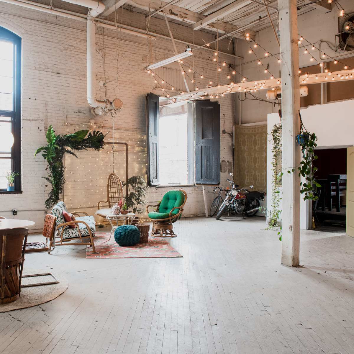 The spacious room features eclectic furniture, including a round table and various seating arrangements, with string lights and plants creating a vibrant atmosphere.