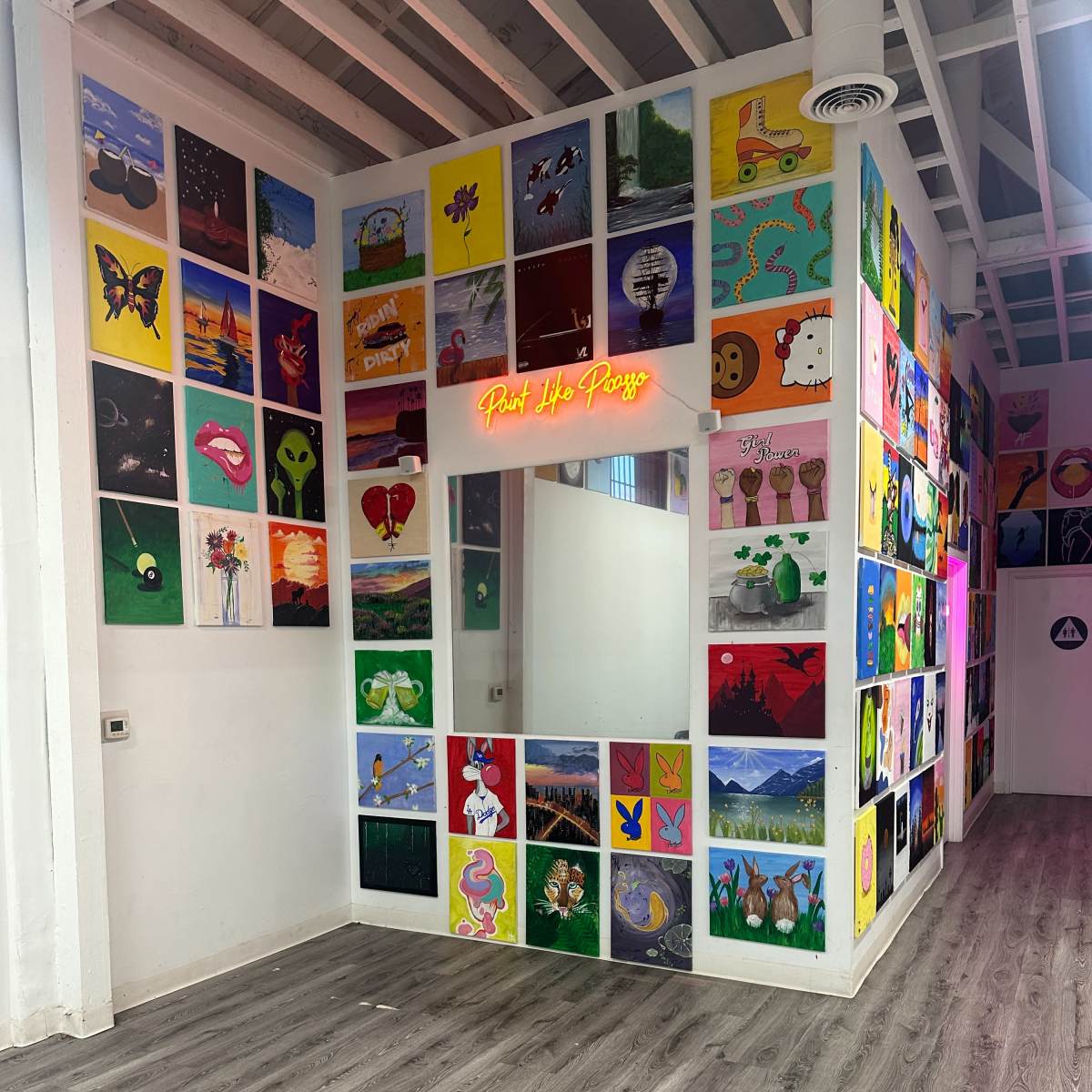 A brightly colored interior room with a wall covered in various artworks, and a mirror framed by neon lights that read "Paint The Party."