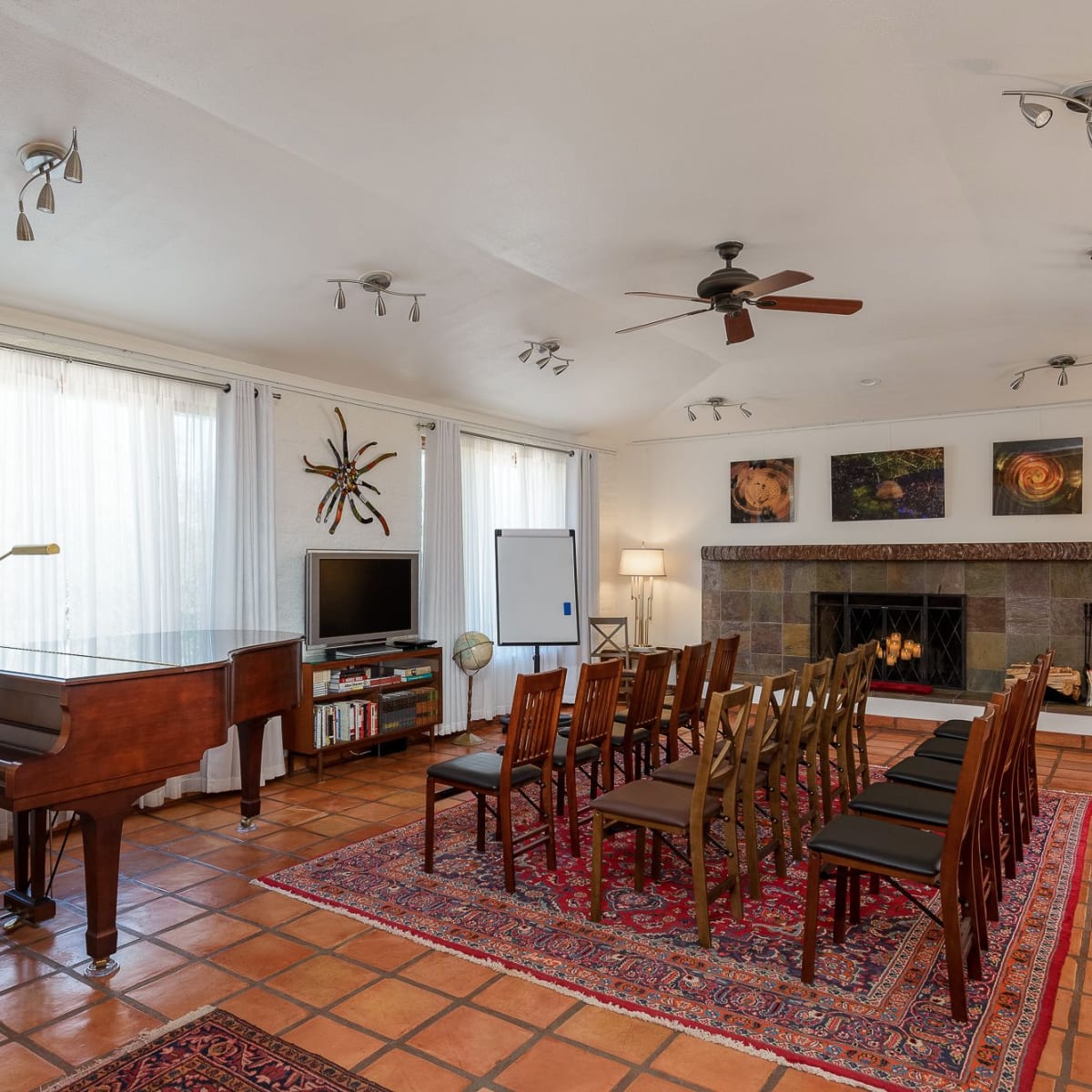 The room features a grand piano, a seating arrangement of wooden chairs, and a fireplace, all surrounded by large windows and decorative wall art.