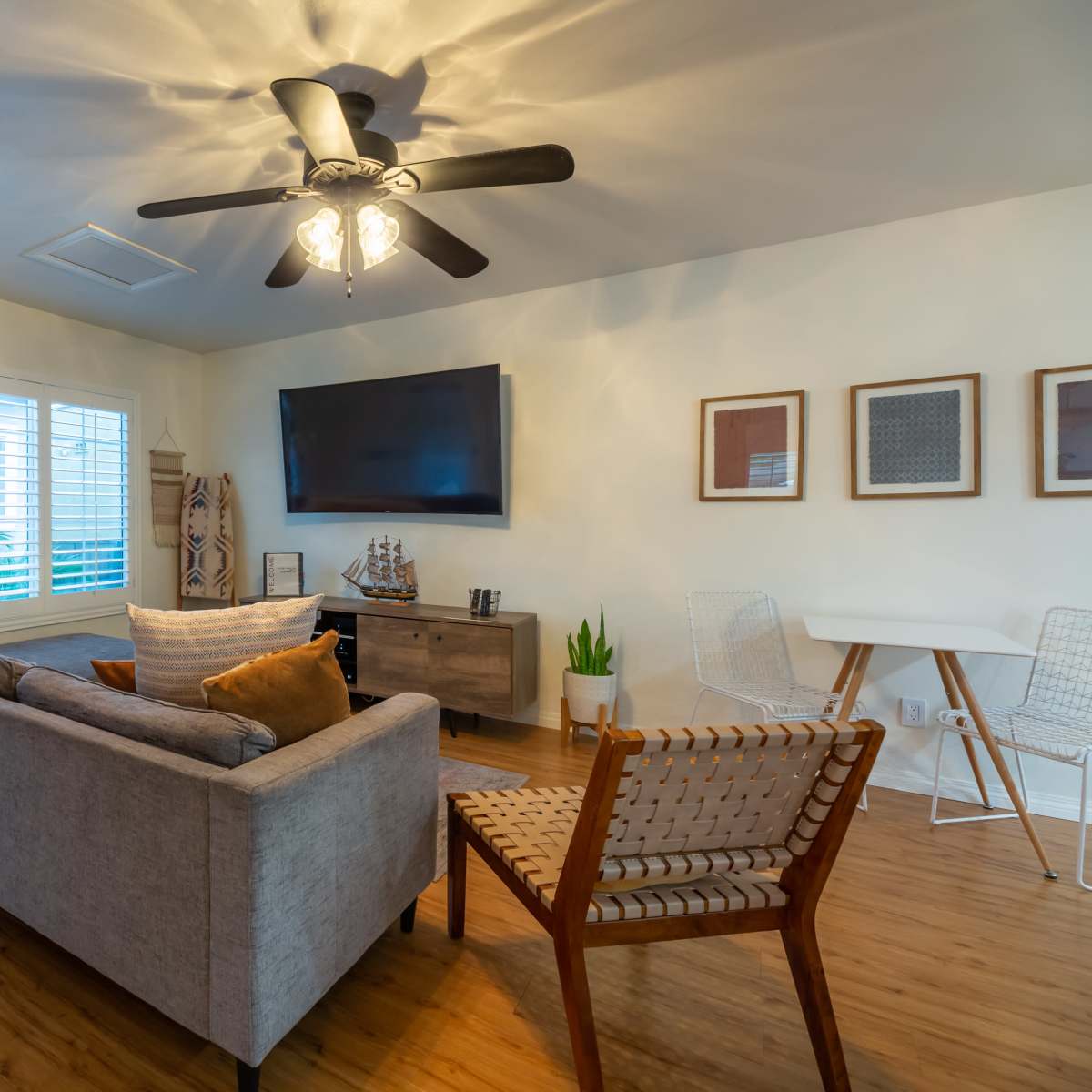 A modern living room features a gray sofa, a wooden television stand, and a ceiling fan, with natural light coming through a side door and windows.