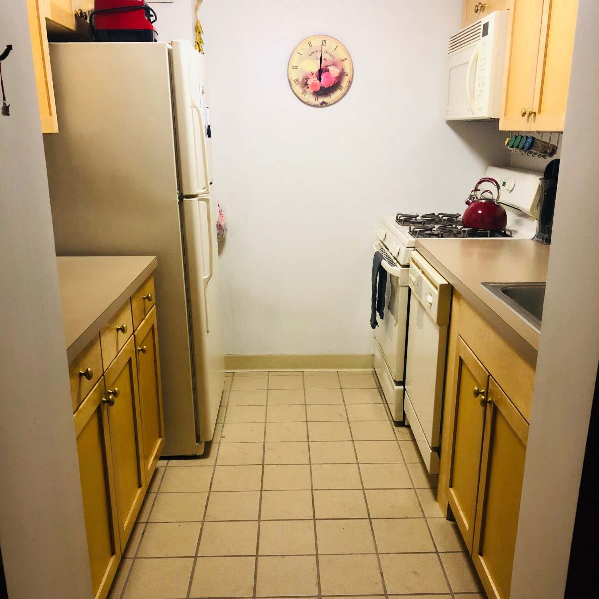A compact kitchen with beige cabinets, a white stove, and a refrigerator, featuring tiled flooring and a wall clock.