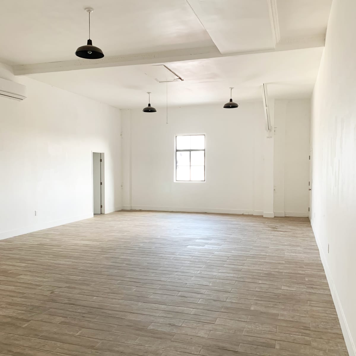 A spacious, empty room with wooden flooring and white walls, featuring a single window and pendant lights.