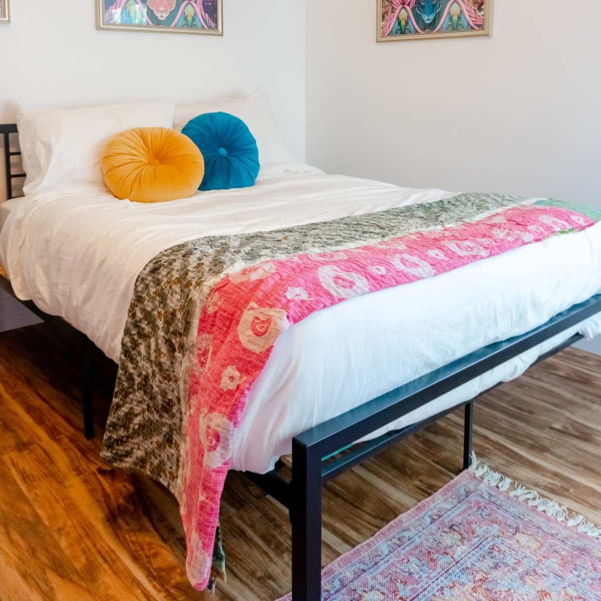 A neatly made bed with colorful decorative pillows and a patterned throw blanket, placed in a well-lit bedroom with two framed artworks on the wall.