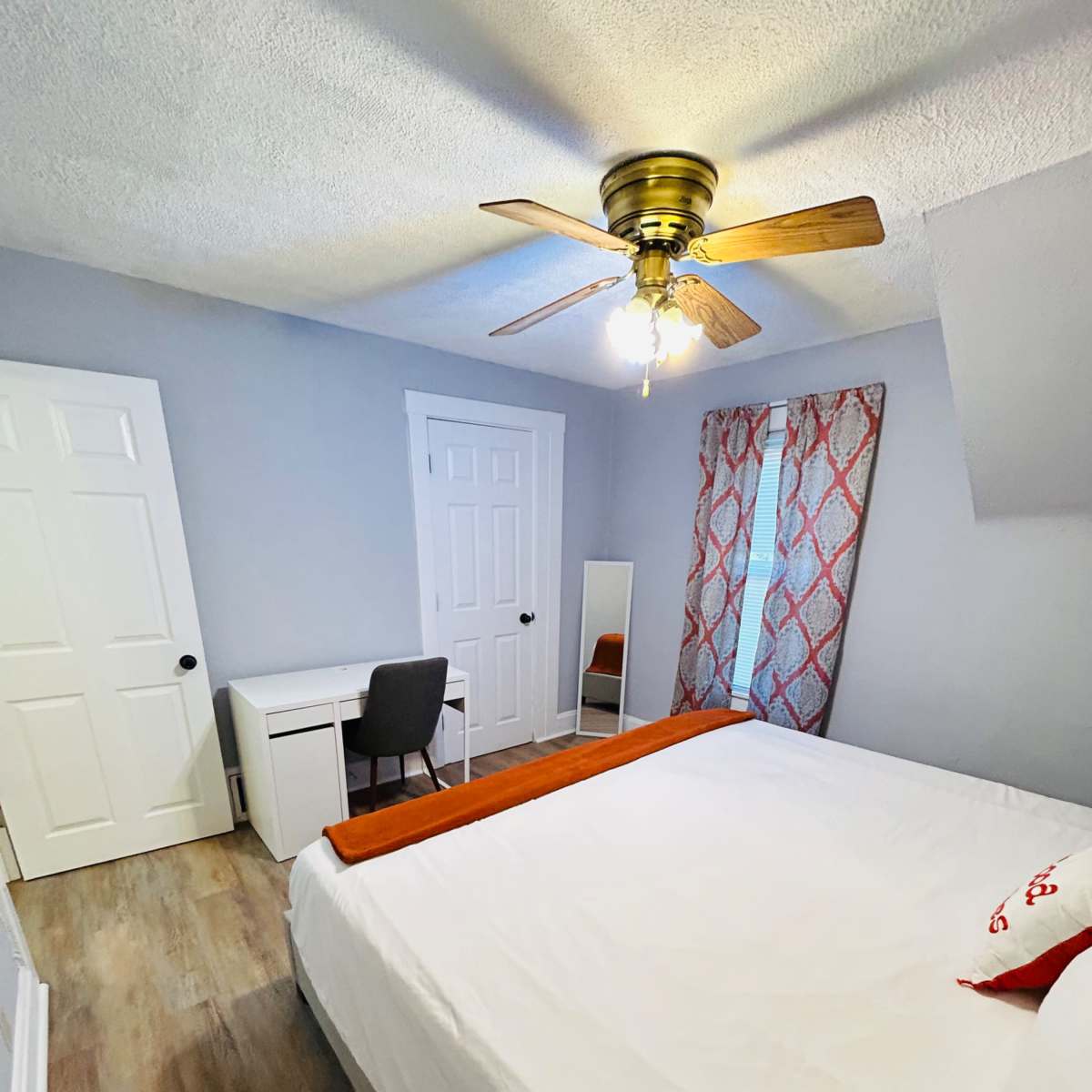 A small bedroom featuring a bed, a desk with a chair, a mirror, a window with patterned curtains, and doors leading to a closet and the outside.