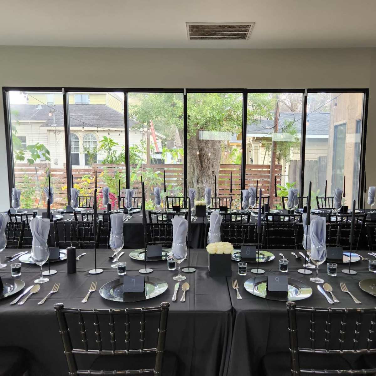 A long dining table is set with black tablecloths, silver cutlery, glassware, and white napkins, facing large windows overlooking a garden.