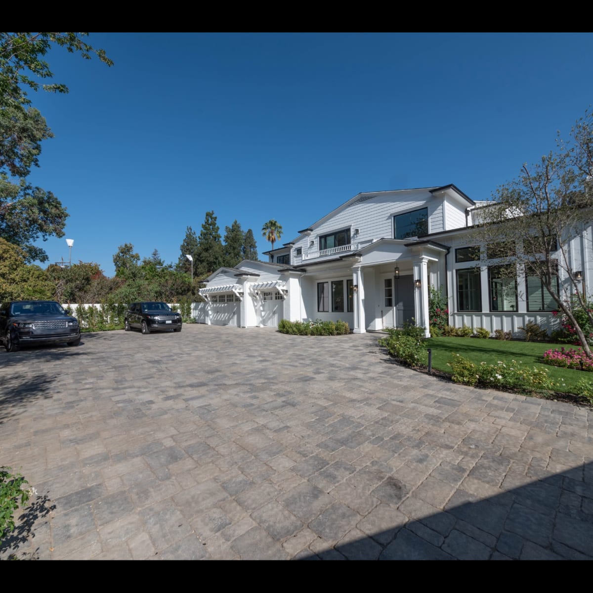 A large modern house with a wide driveway made of pavers, surrounded by lush greenery and landscaping under a clear sky.