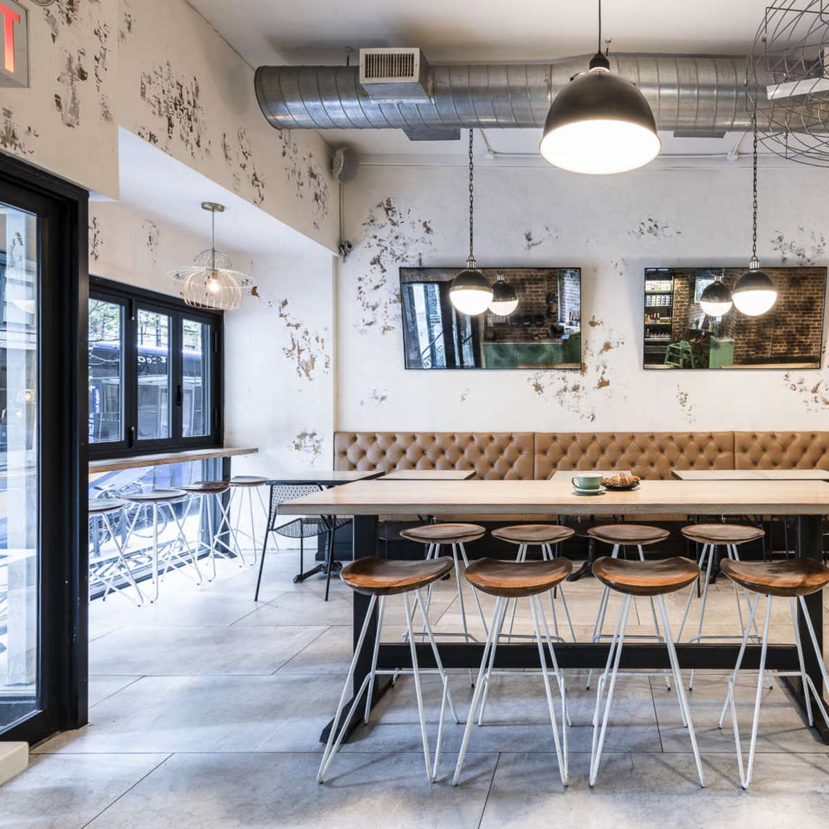 A modern café interior featuring a long wooden table with stools, decorative wall mirrors, and large windows allowing natural light.