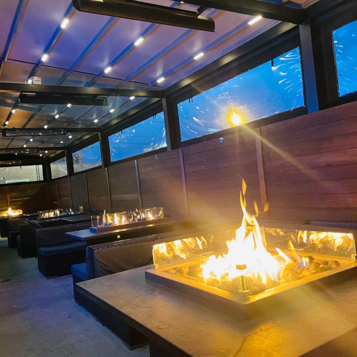 A modern, covered outdoor seating area with several fire pits and a view of a rainy evening outside.