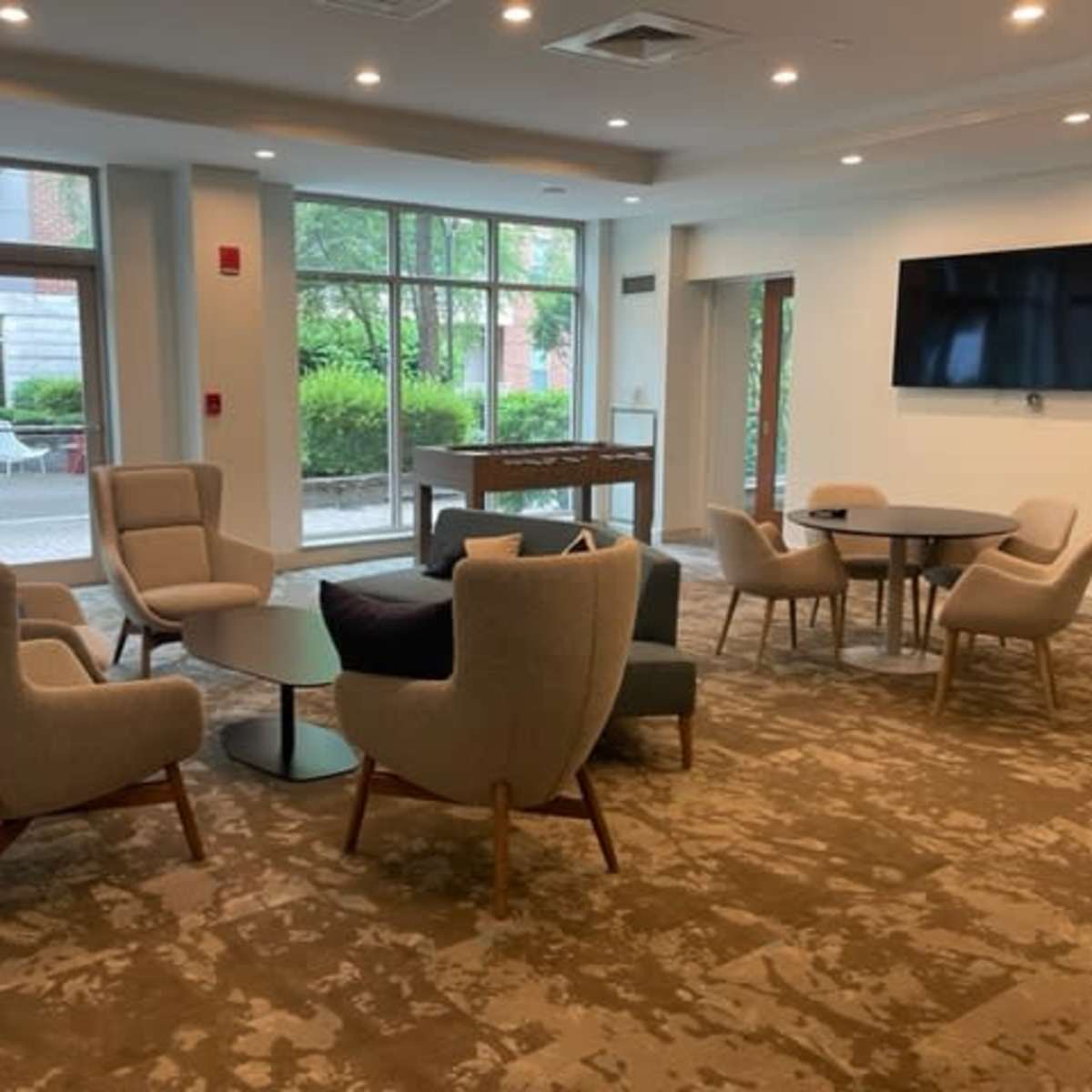 A modern lounge area features several upholstered chairs, a coffee table, and a large wall-mounted television, with windows overlooking a green outdoor space.