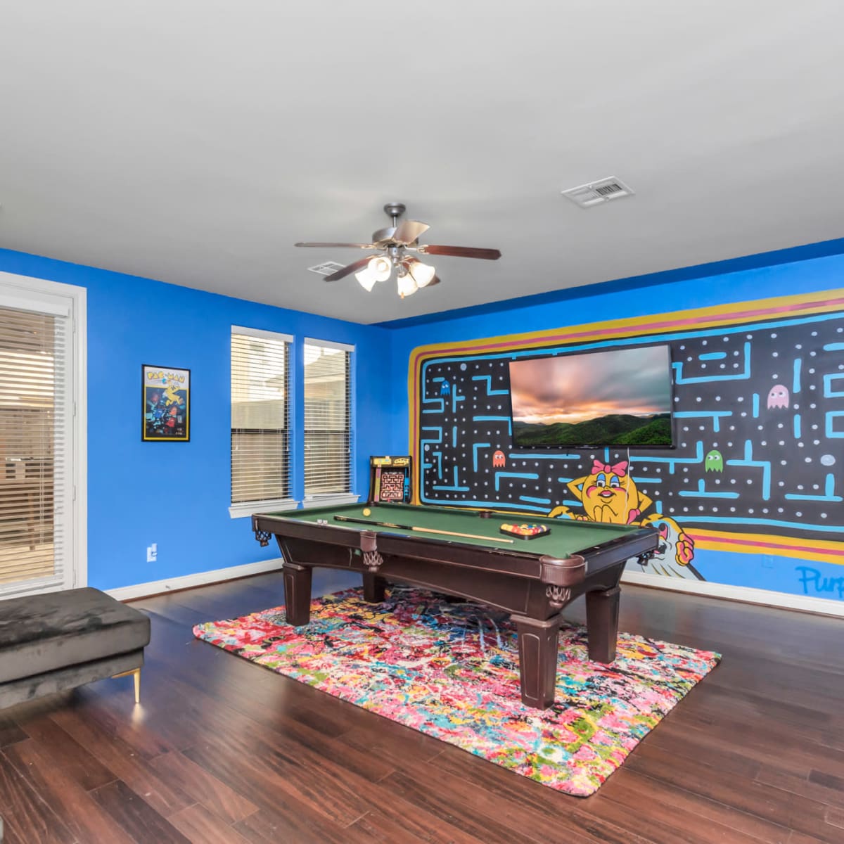 A game room with a pool table, a colorful rug, a large television on the wall, and blue painted walls featuring a mural.