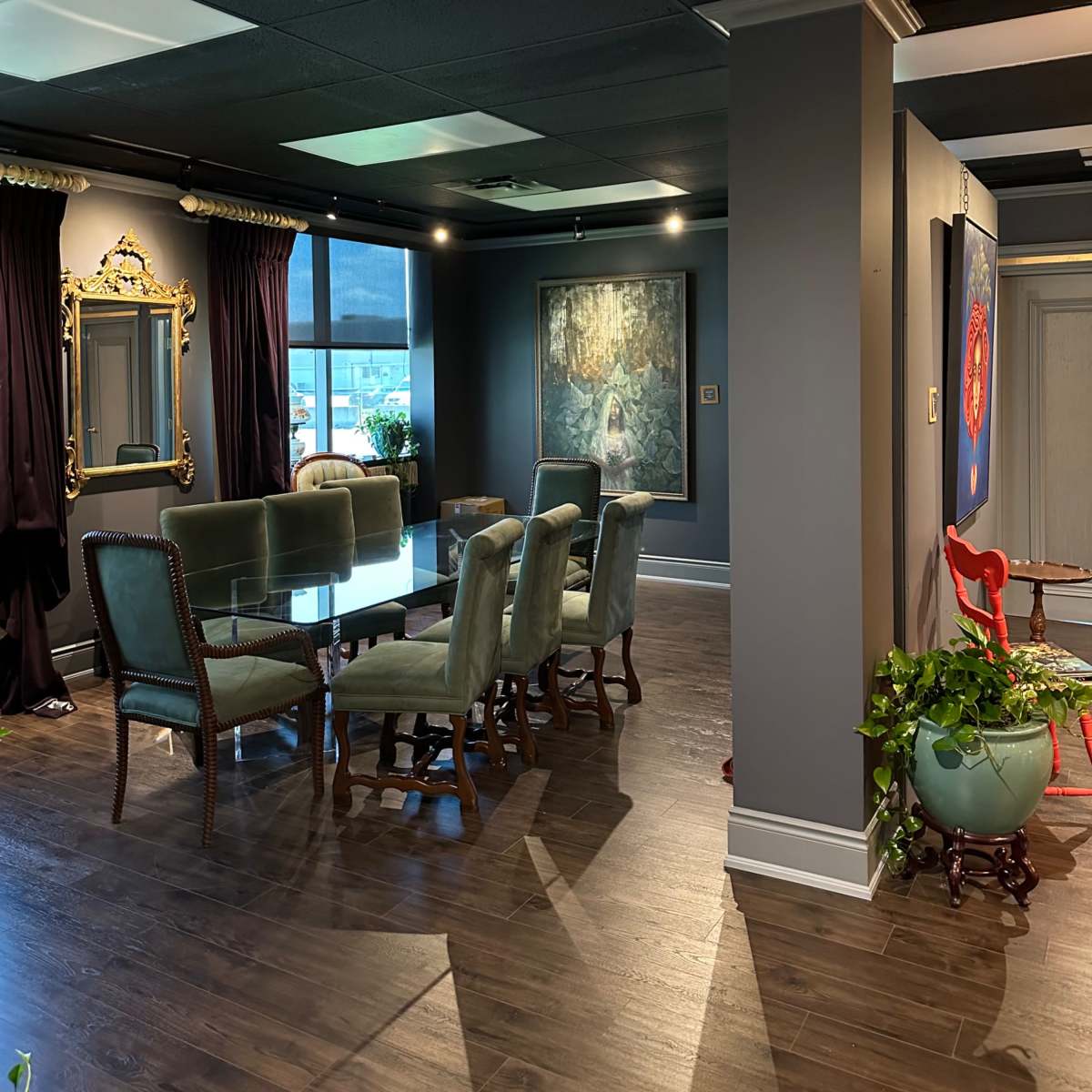 A modern interior space features a dining table surrounded by green chairs, artwork on the walls, and large windows with drapes.