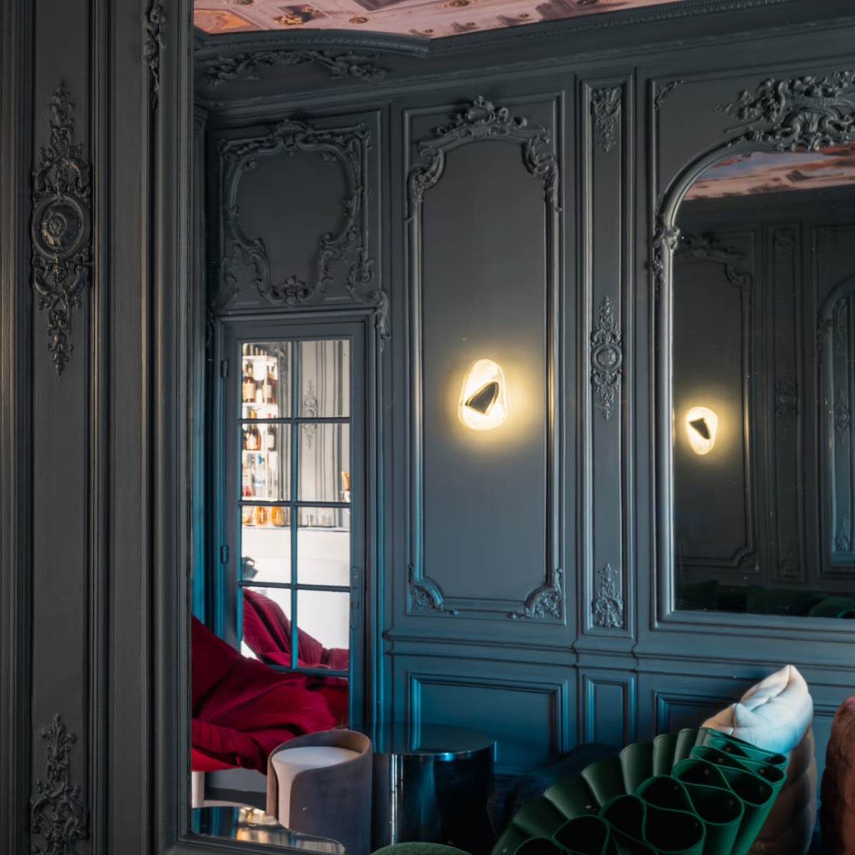 An elegantly decorated room with ornate green walls, illuminated wall sconces, and a reflection in a large mirror.