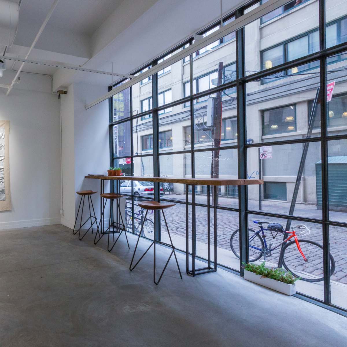 An art gallery features a textured American flag artwork on the wall beside a minimalist seating area with a long table and a bicycle visible outside through large windows.