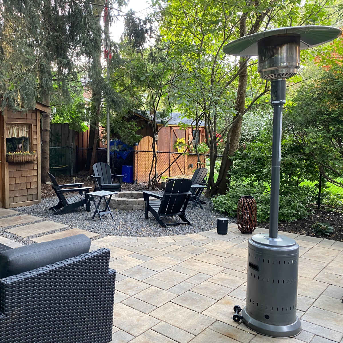 A cozy outdoor patio features a seating area with black furniture, a fire pit, and a patio heater surrounded by trees and a shed.