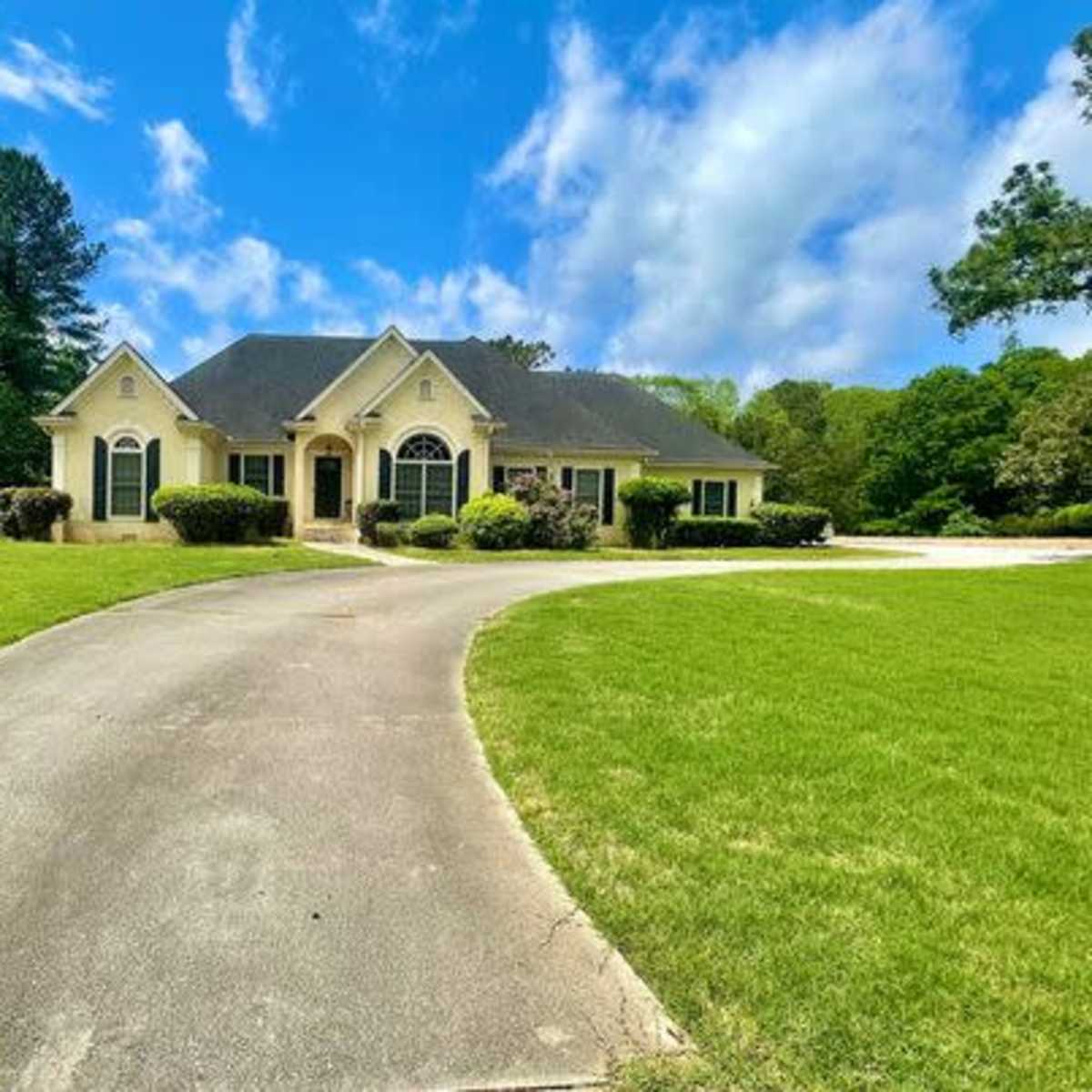 A large, two-story house with a neatly landscaped yard sits at the end of a curved driveway under a blue sky.