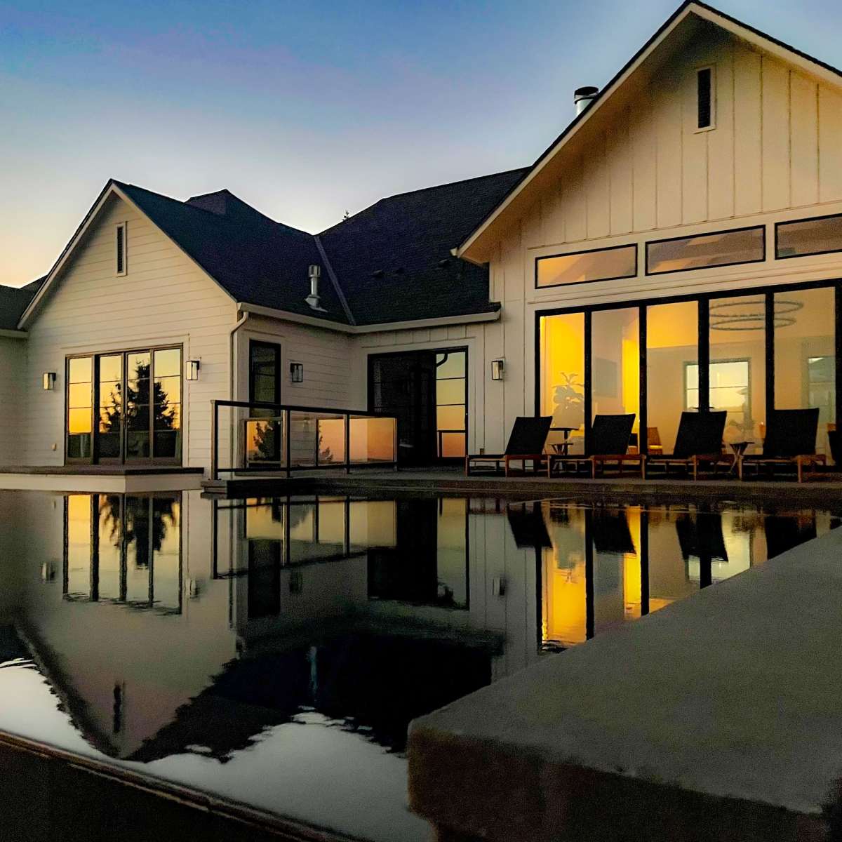 A modern house with large windows reflects in a calm pool at sunset.