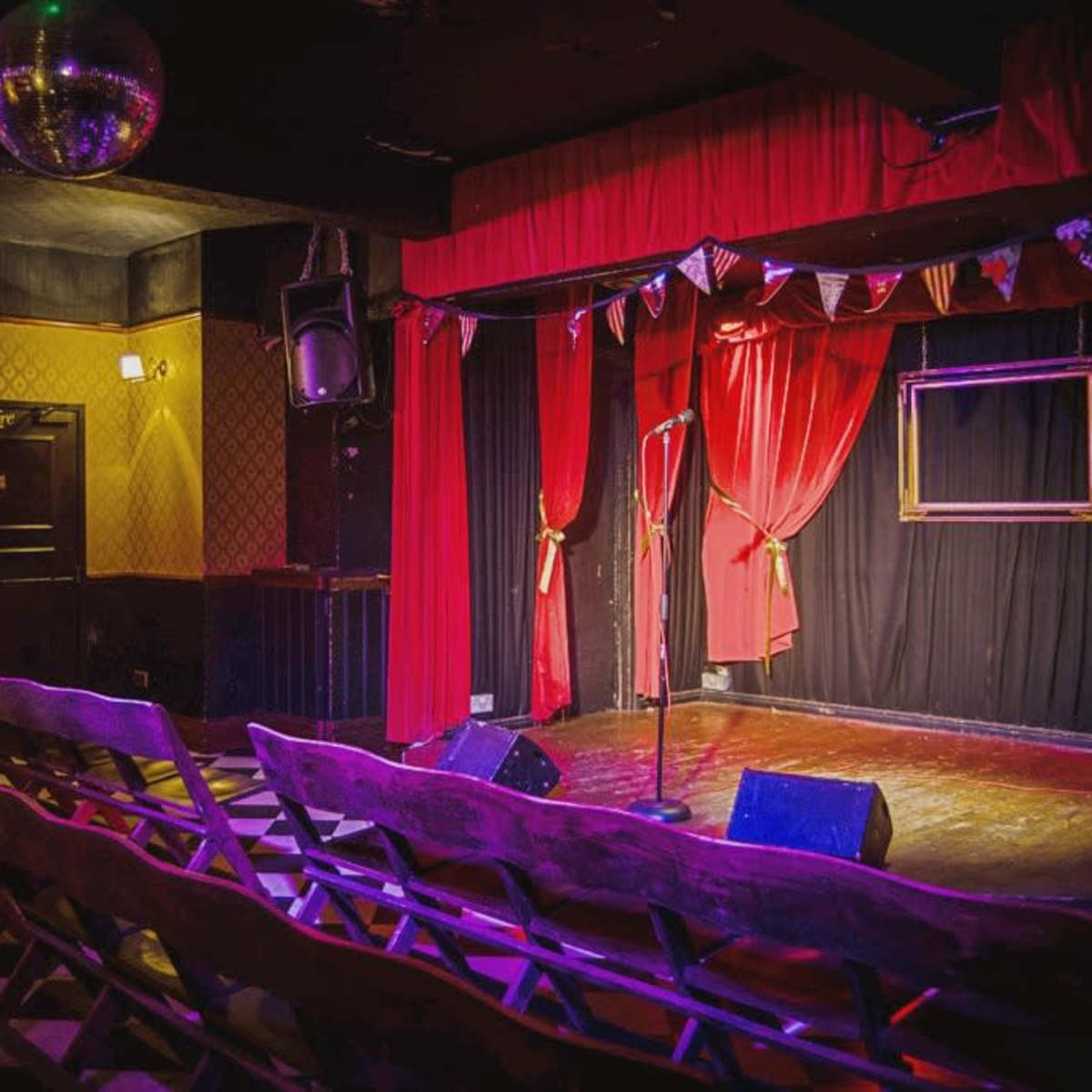 A small stage with a microphone stand, surrounded by red curtains and empty wooden benches facing the performance area, illuminated by colorful lights and a disco ball hanging from the ceiling.