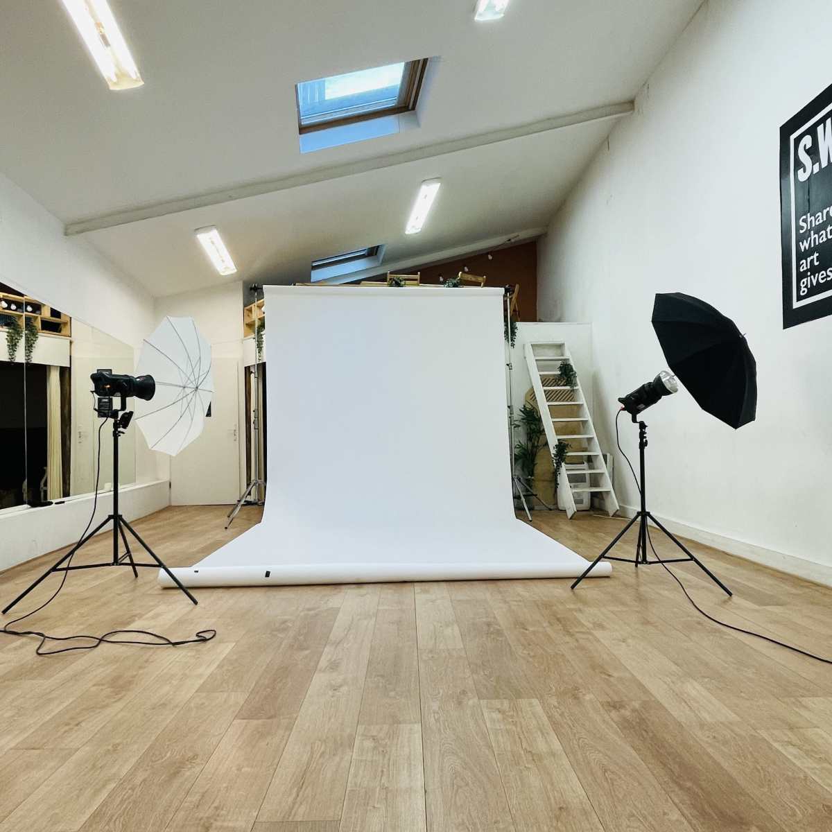 A photography studio features a white backdrop with lighting equipment set up on either side, along with a staircase and mirrors in the background.