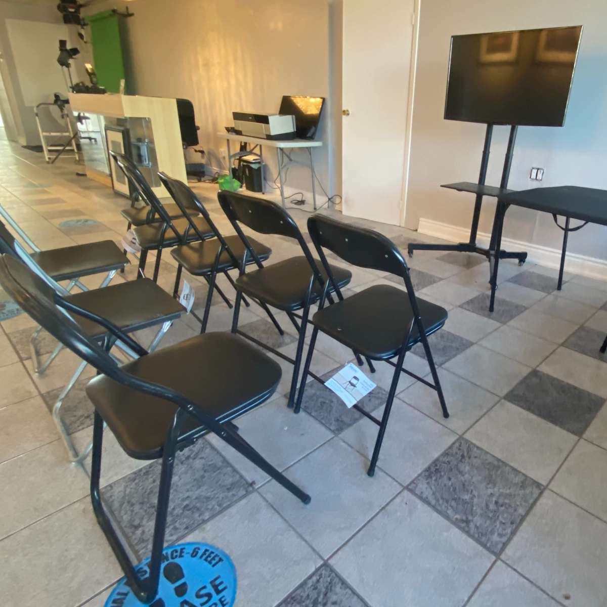A room with a row of black folding chairs facing a television on a table, with a computer station visible in the background.