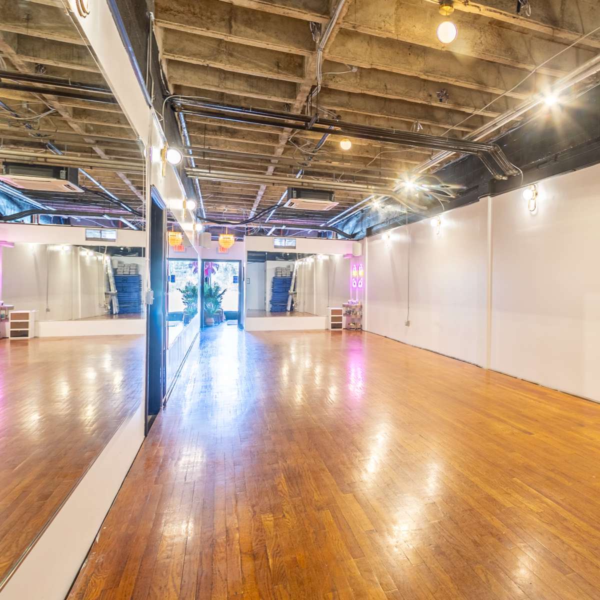 A spacious indoor room with polished wooden floors, large mirrors on one wall, and fitness mats stacked against another wall.