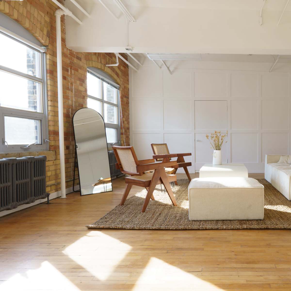 A bright and spacious living room with a beige sofa, a matching coffee table, two wooden chairs, a mirror, and a large area rug on a hardwood floor.