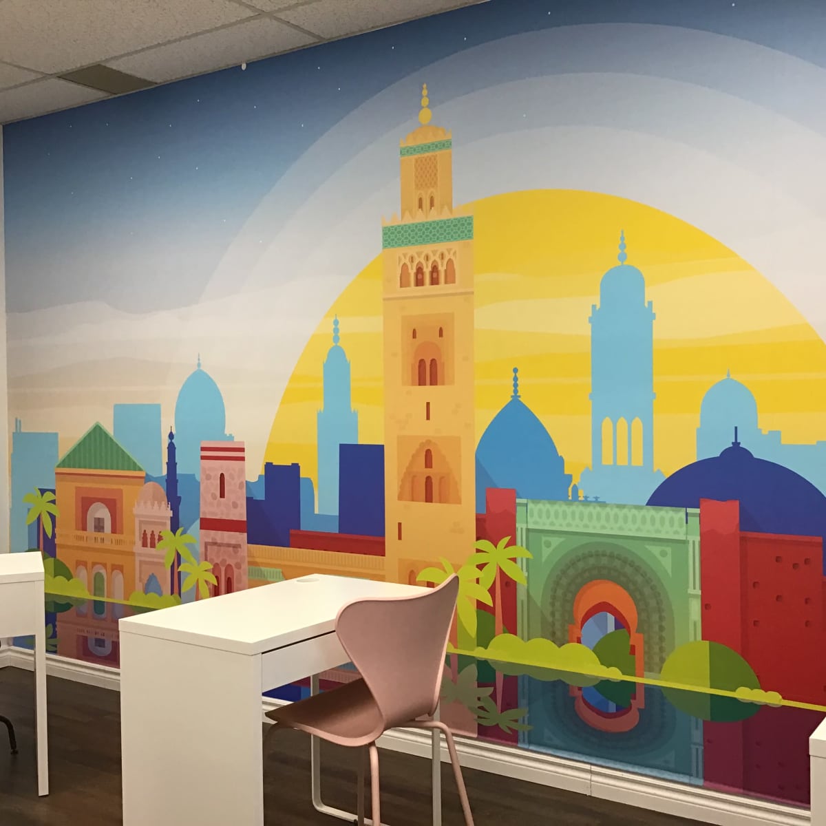 A brightly colored mural featuring a skyline with various architectural styles, set against a large yellow sun, in a room with white desks and a pink chair.