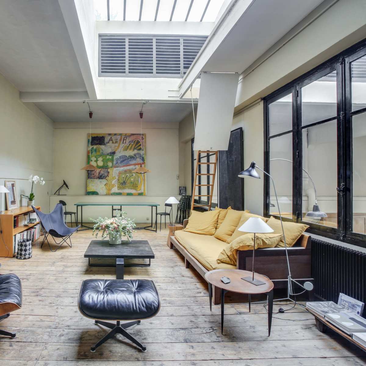 A spacious, modern living room with wooden flooring, large windows, a yellow sofa, and various pieces of furniture including an armchair and a coffee table.
