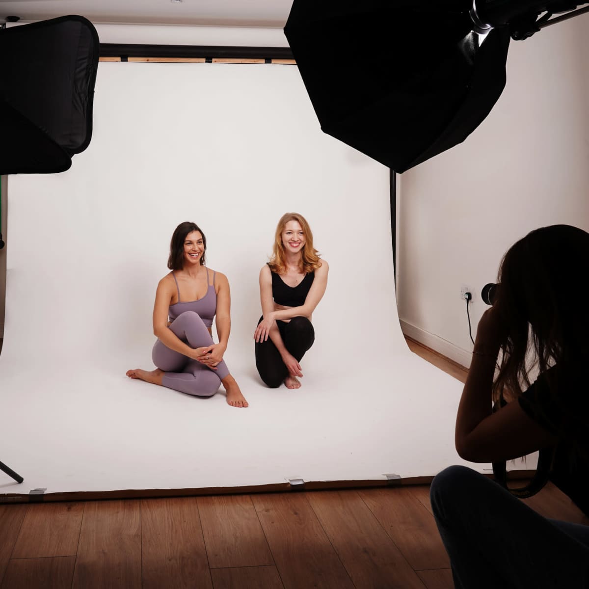 Two women pose seated on a backdrop while a photographer captures their image in a studio.