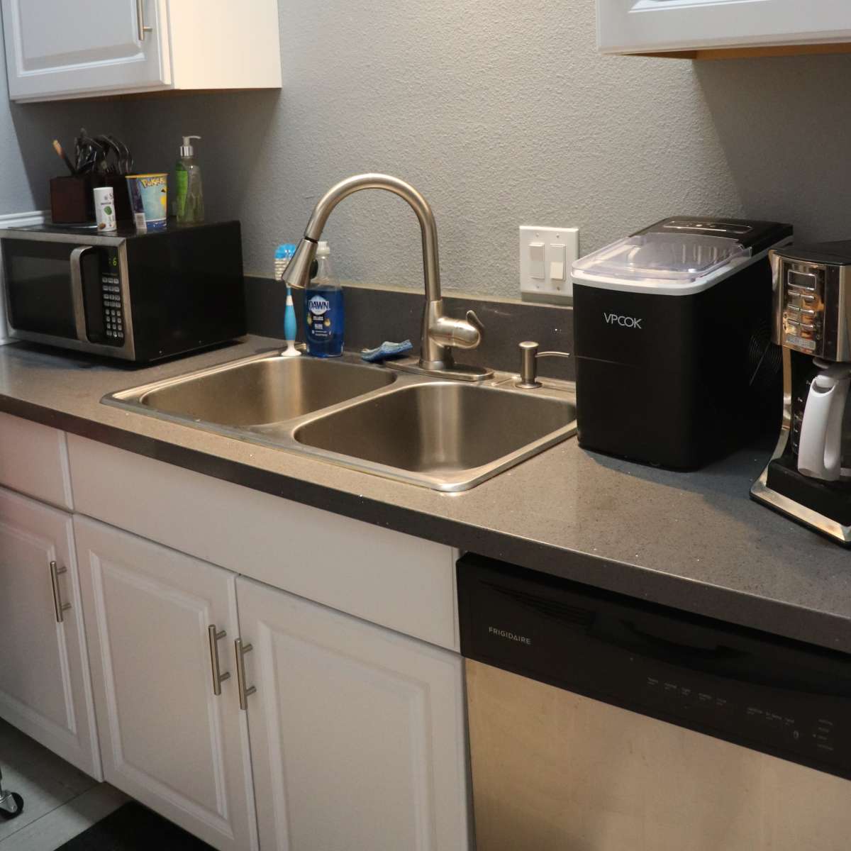 A kitchen sink area with two stainless steel sinks, a microwave, a countertop appliance, and a dishwasher beneath white cabinets.