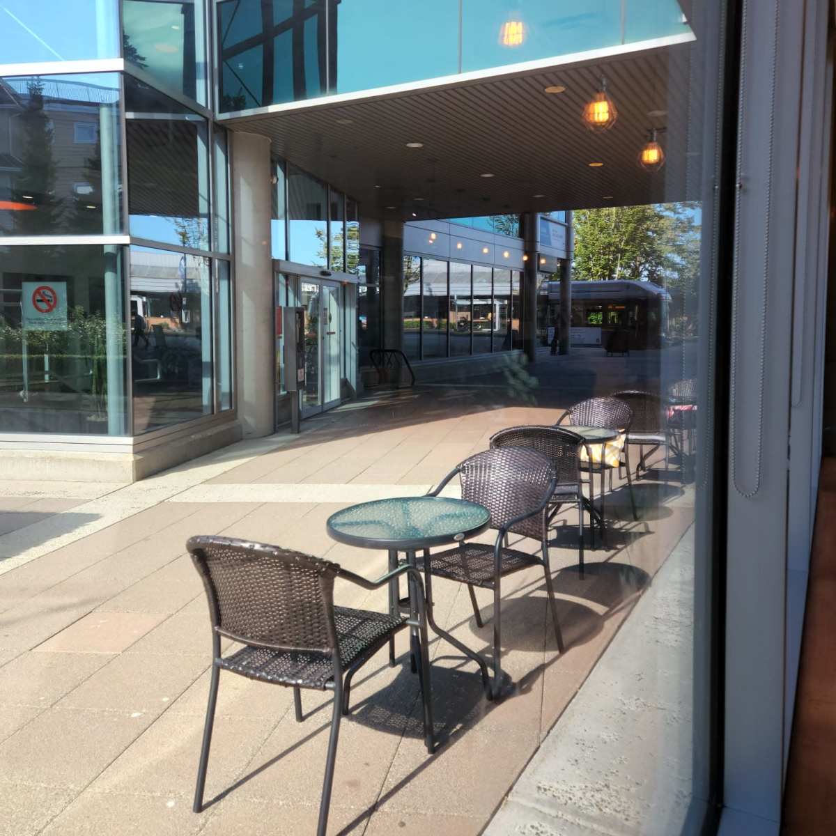 An outdoor seating area with empty tables and chairs outside a modern building, reflecting bright sunlight.
