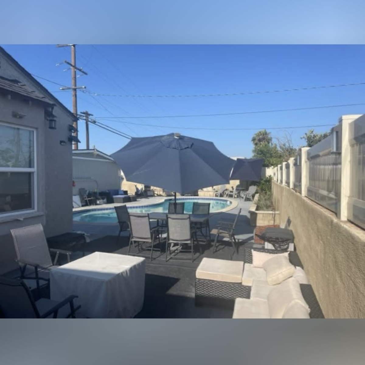 A pool area is visible with lounging chairs, tables, and large umbrellas, surrounded by a fence and a clear blue sky.