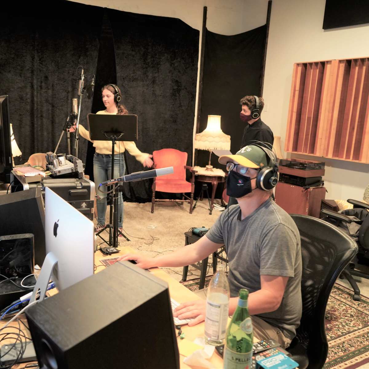 A seated person works at a computer while two others record vocals in a sound studio.